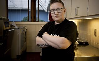 Portræt af Kaja Larsen, der står med sort t-shirt og korslagte arme i et køkken. 